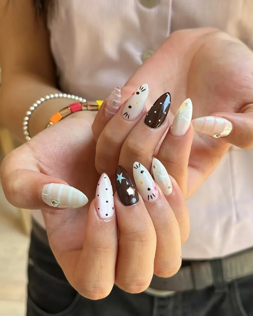 Neutral Hello Kitty nails in brown, cream, and white with hand-drawn Kitty faces, pearls, stripes, and stars on almond nails