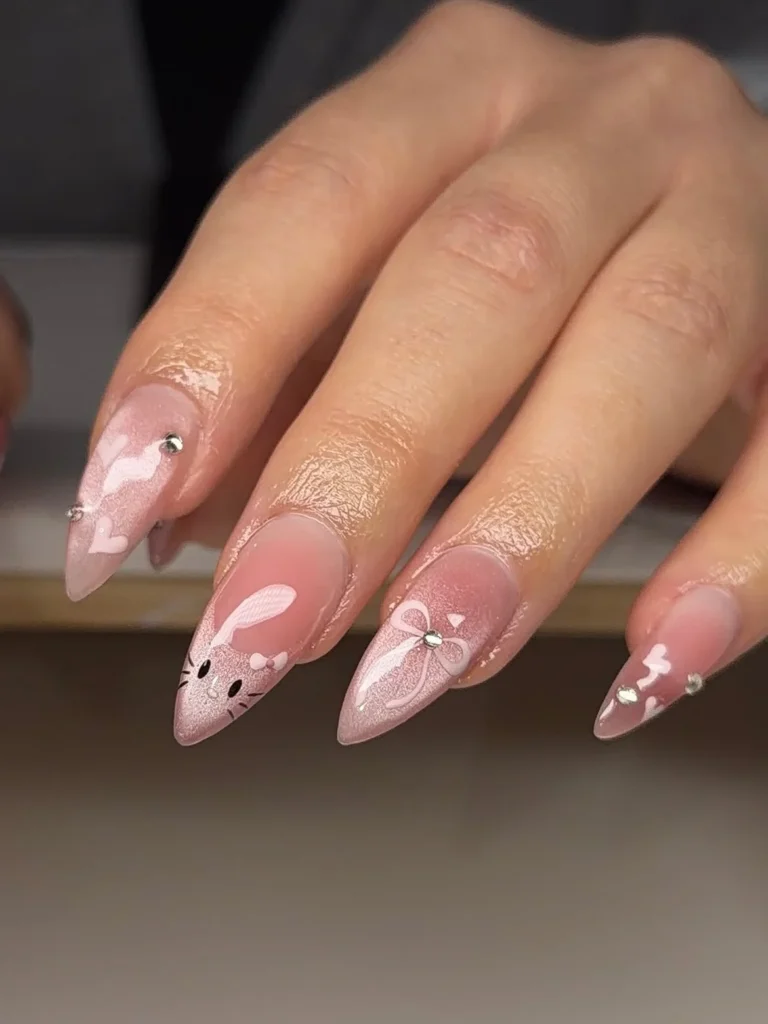 Soft pink cat eye Hello Kitty nails with white freehand Kitty face, bows, hearts, and small rhinestones on long almond nails