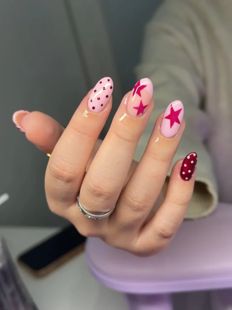 Cool girl nail designs with soft pink base and cherry red stars, dots, and burgundy accent on almond nails